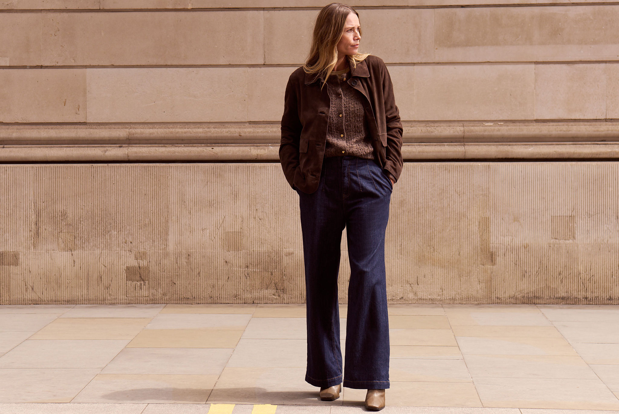 Person in brown jacket and blue wide-leg trousers standing on the pavement in front of a stone wall.